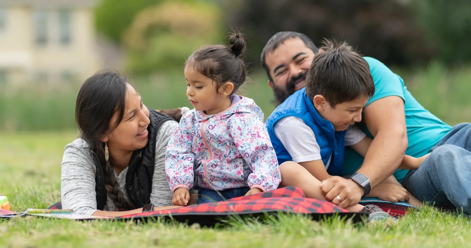 Family of four sitting on a blanket in the grass reading and laughing
